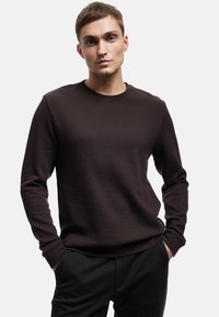 Bruine textuur trui met ronde hals en lange mouwen, voorzien van een subtiel ribbelpatroon. Gecombineerd met een donkere broek poseert het model met de handen in de zakken.