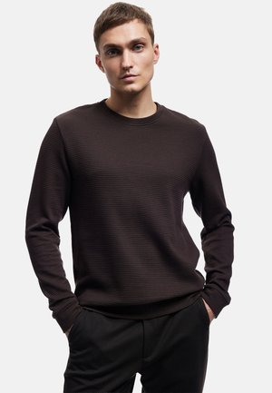 Sudadera de cuello redondo texturizada en marrón con mangas largas, que presenta un sutil patrón de canalé. Combinada con pantalones oscuros, el modelo posa con las manos en los bolsillos.