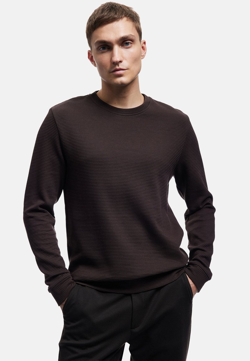 Bruine textuur trui met ronde hals en lange mouwen, voorzien van een subtiel ribbelpatroon. Gecombineerd met een donkere broek poseert het model met de handen in de zakken.