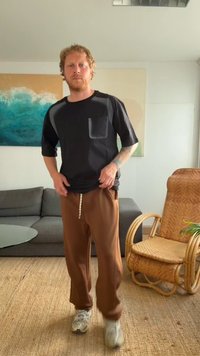 Μαύρο κοντομάνικο πουκάμισο με τσέπη, συνδυασμένο με καφέ φαρδιά παντελόνια. Το σκηνικό περιλαμβάνει ένα γκρι καναπέ και μια πλεκτή καρέκλα.