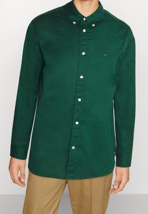 Homme portant une chemise verte foncée boutonnée avec un petit logo sur la poitrine, associée à un pantalon beige, debout devant un fond uni.