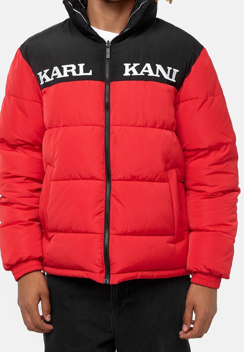 Personne portant une doudoune rouge et noire avec "KARL KANI" en lettres blanches sur la poitrine, fermeture éclair fermée, debout avec les bras détendus.
