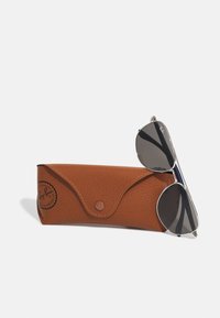 Occhiali da sole Ray-Ban aviator con lenti grigie scure e montatura argento, appoggiati accanto a un astuccio marrone testurizzato con chiusura a scatto e logo.