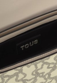 Beżowa skórzana torba w środku z czarnym wykończeniem, z logo "TOUS" w kolorze srebrnym oraz kremowym zewnętrzem ozdobionym delikatnym wzorem kwiatowym.