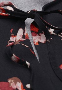 Sort blomstret stof med røde og pink mønstre. Har en bindebånd ved halsudskæringen og en blød, glat tekstur. Nærbillede af designet.