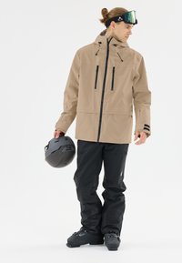Beige wasserdichte Jacke mit schwarzen Reißverschlüssen, zwei Brusttaschen und verstellbarer Kapuze, kombiniert mit schwarzen Skihosen und einem grauen Helm.