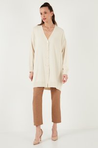 Kvinde iført en lang cremefarvet cardigan med knapper, brune korte bukser, beige højhælede sko og lagdelte halskæder, stående foran en ensfarvet baggrund.