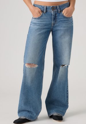 Personne portant un jean bleu taille haute à jambes larges avec des déchirures aux deux genoux et des chaussures noires, mains dans les poches avant.