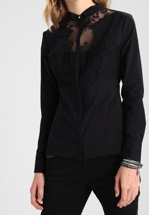 Vrouw draagt een nauwsluitende zwarte blouse met kantdetails op de borst en schouders, gecombineerd met een zwarte broek en zilveren armbanden.