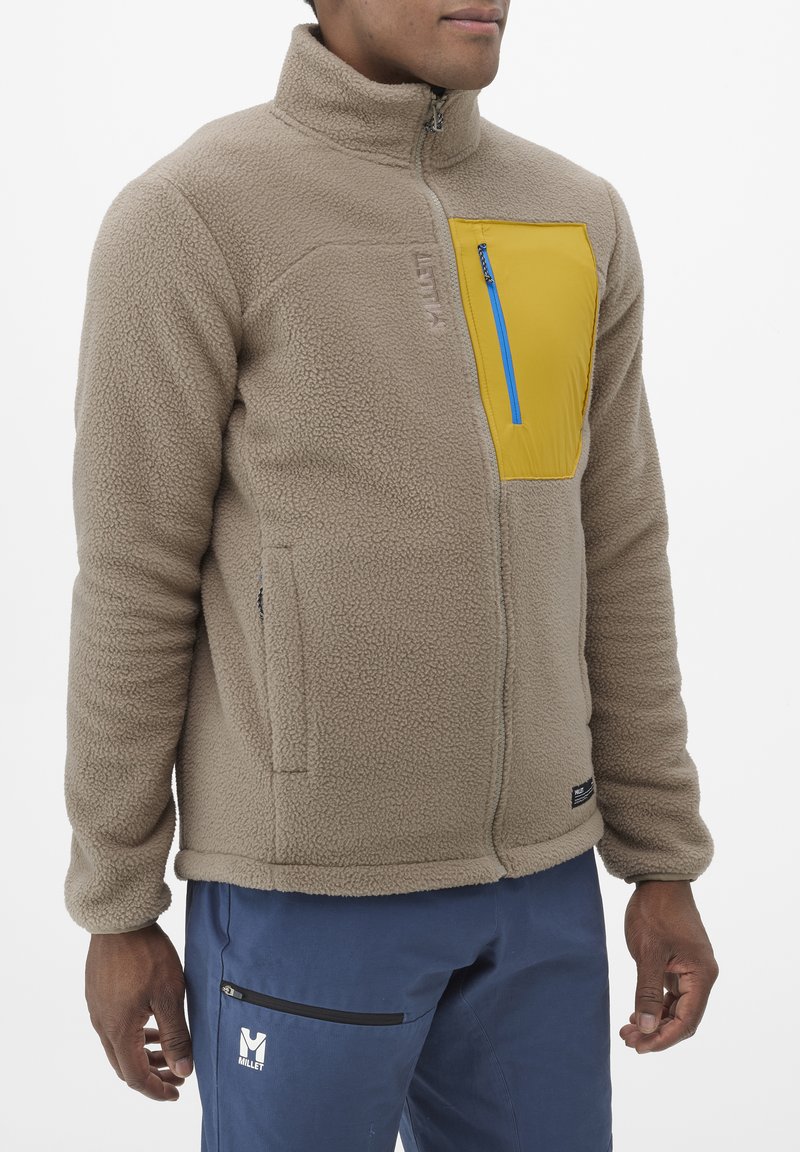 Veste en polaire beige avec col montant, poche poitrine zippée jaune avec zip bleu, poches latérales et détail en tissu texturé.