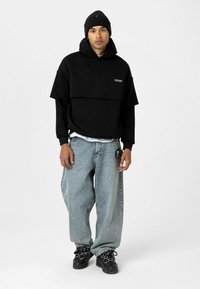 Sweatshirt à capuche oversize noir superposé sur une chemise blanche, associé à un jean en denim bleu ample et des baskets noires.