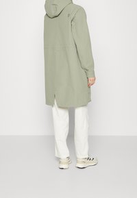 Veste à capuche vert clair à coupe droite, avec une fente au dos et un logo brodé ; associée à un pantalon blanc et des baskets beiges.