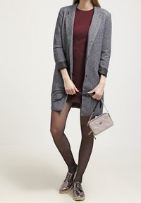 Abrigo texturizado gris sobre un vestido burdeos, combinado con medias negras transparentes y zapatos oxford metálicos brillantes. Se sostiene un pequeño bolso acolchado.