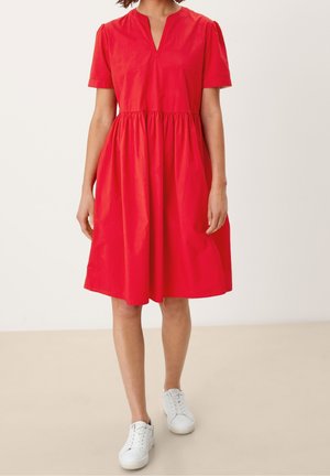 Mujer vestida con vestido rojo hasta la rodilla con mangas cortas y escote en V, combinado con zapatillas blancas, de pie sobre suelo beige y fondo blanco.