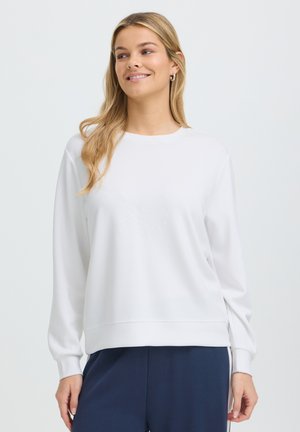 Femme blonde portant des boucles d’oreilles créoles dorées, un pull blanc à manches longues et un pantalon bleu marine, souriante et regardant sur le côté.