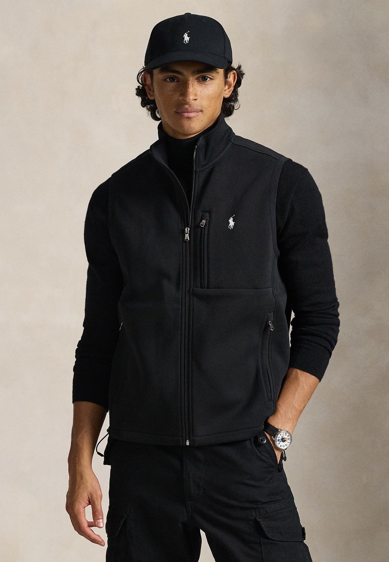 Μαύρο γιλέκο από φλις με φερμουάρ, μπροστινή τσέπη και μικρό λογότυπο. Φορεμένο πάνω από μαύρο πουλόβερ, συνδυασμένο με αντίστοιχο μαύρο καπέλο.