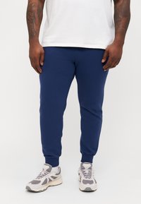 Marineblå joggebukser i et mykt materiale med en tettsittende design, ribbede mansjetter og glatt tekstur, kombinert med lysegrå joggesko.