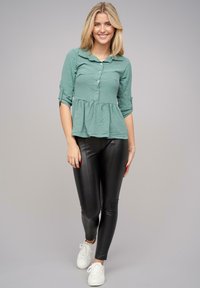 Top peplum verde, con bottoni e colletto, maniche a tre quarti, abbinato a leggings neri lucidi e sneakers bianche. Texture semplice.