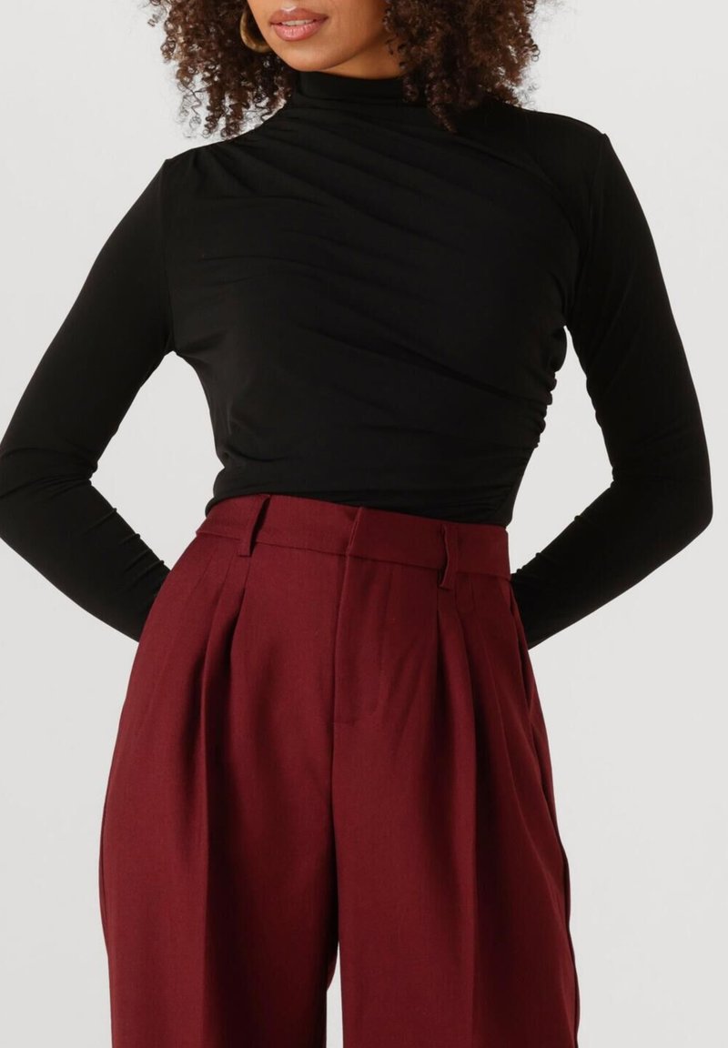 Vrouw met krullend haar, gekleed in een fitted zwarte lange mouw turtleneck en hooggetailleerde wijde donkerrode broek tegen een witte achtergrond.