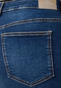 Jeans de ganga azul escura com uma superfície texturizada e suave. Apresenta um bolso traseiro e costura contrastante ao longo das bordas. Rótulo visível na cintura.