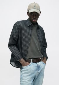 Tumesinises ruudulises nööpidega särgis kihiliselt üle tumerohelise T-särgi. Hele sinised teksapüksid, pruun vöö ja beež lihtsa disainiga nokamüts.