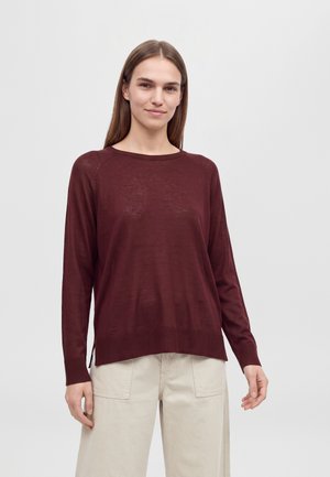Un mannequin aux longs cheveux bruns porte un pull bordeaux à manches longues et un pantalon crème sur un fond uni et clair.