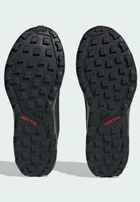 Suela de zapatos de exterior negra con profundos surcos de tracción. Presenta goma moldeada con los logotipos "TRAXION" y "adidas" en rojo y blanco.