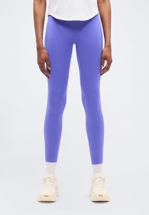 Leggings violets en tissu extensible, avec une texture lisse et élégante et une taille haute. Associés à des chaussures de sport de couleur claire.