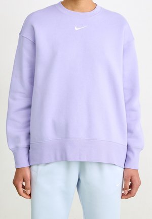Personne portant un sweat-shirt Nike violet clair et un pantalon bleu clair avec des logos Nike, debout, les mains détendues le long du corps.