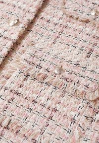 Rosa- und cremefarbenes gewebtes Stoffmaterial mit ausgefransten Kanten und schwarzen Akzenten. Verfügt über zwei dekorative Metallknöpfe und eine strukturierte Oberfläche.