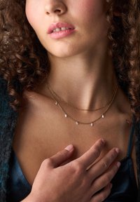 Collana a strati dorata con accenti a forma di marquise, posizionata su una clavicola scoperta. La mano della modella poggia delicatamente sul suo petto.