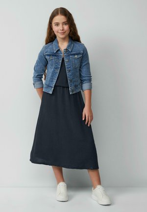 Veste en denim au design ajusté, couleur bleue, avec des accessoires en métal argenté, portée sur une robe maxi texturée gris foncé, accompagnée de baskets blanches.