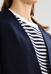 Marinblå blazer med en mjuk textur, lager på lager över en vit och marinblå randig skjorta. Designen har en enkel halsringning och rena linjer.