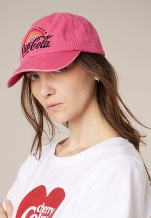Jeune femme portant une casquette Coca-Cola rose délavée avec un arc-en-ciel et un t-shirt blanc avec un logo Cherry Coke rouge.