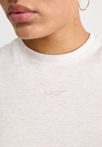 Light gray ribbed Nike t-shirt featuring a small beige embroidered logo on the chest, worn by a person with a heart tattoo on the neck.