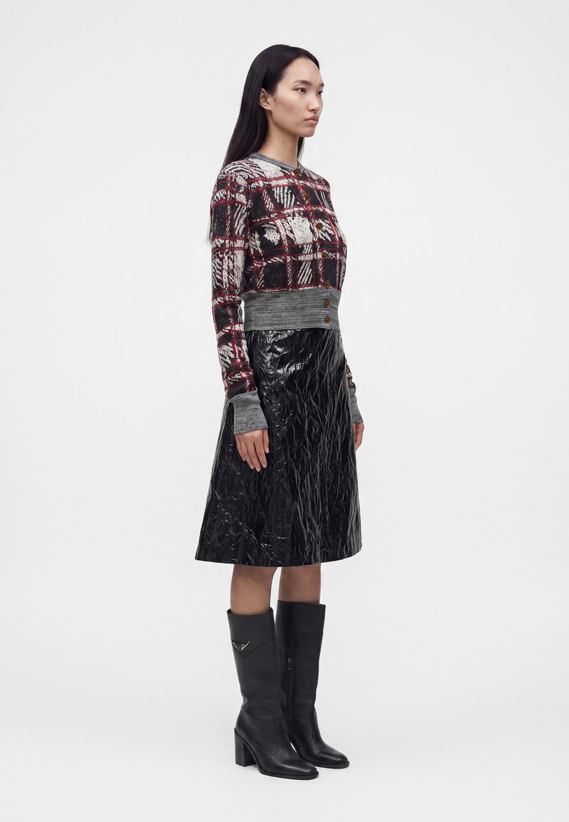 Femme portant un cardigan à boutons à motifs avec des poignets gris, une jupe midi noire brillante et des bottes en cuir noires montant jusqu'aux genoux, debout de profil.