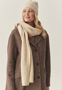 Beige gebreide muts en bijpassende sjaal om de nek gedrapeerd, gecombineerd met een visgraatmantel in bruin- en roesttinten, voorzien van grote knopen.