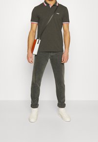 Polo gris foncé avec bordure de col rouge et blanche, associé à un jean gris et des baskets blanches. Le mannequin tient un sac à bandoulière bicolore.