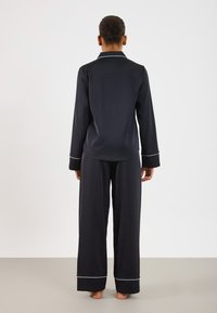 Svart sateng pysjamassett med lange ermer, en kragekjortel med hvite kanter, og vide bukser. Smooth tekstur, avslappet passform.