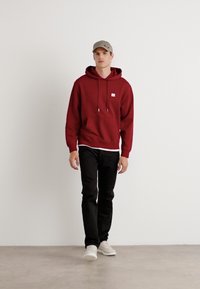 Felpa rossa con cappuccio e cordini, con patch logo sul petto, indossata sopra una maglietta bianca, con jeans neri e sneakers chiare, su uno sfondo semplice.