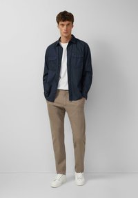 Marineblaues Hemd mit Knöpfen und zwei Brusttaschen, getragen über einem weißen T-Shirt. Hellbraune Hose und weiße Sneakers vervollständigen das Outfit.