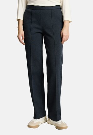 Frau trägt marineblaue Hosen mit geradem Bein, cremefarbenes Langarmshirt und weiße Sneakers mit beigen Sohlen, steht vor einem einfarbigen Hintergrund.