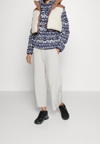 Maglione in pile a fantasia navale sotto un gilet imbottito beige, abbinato a pantaloni plissettati grigio chiaro e scarpe da ginnastica nere.