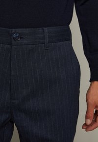 Pantalon bleu marine foncé à fines rayures avec fermeture à boutons, devant plat et tissu texturé. Comprend une poche latérale et des passants pour ceinture.