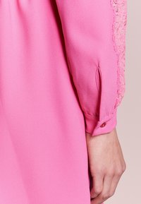 Rosa blus med en slät yta, med spetsdetaljer på ärmarna. Manschetten har en knappdetalj och en liten spricka för rörelsefrihet.