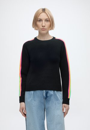 Woman with short blonde hair wearing a black sweater with rainbow stripes on sleeves and light blue high-waisted jeans standing against white background.