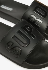 Sorte slidssandaler med en bred rem med præget "SUP" bogstaver, glat syntetisk materiale og en tekstureret ydersål for godt greb.
