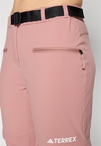 Roze kratke hlače za vanjsku upotrebu s crnim podesivim remenom, koje imaju dva džepa sa zatvaračem i bijeli logo na donjem rubu. Lagani materijal, glatka tekstura.