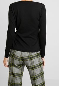 Černý dlouhý rukáv top s kulatým výstřihem, spárovaný se šedými kostkovanými kalhotami s žlutými a zelenými pruhy, viditelnými na zadní straně.
