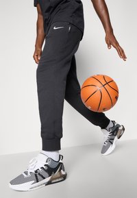 Melnas treniņbikses ar baltu Nike logo, apvienotas ar melnbaltiem sporta apaviem, kas aprīkoti ar sietu paneļiem un gaisa zoli, blakus oranžai basketbola bumbai.